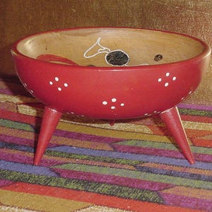 May include: A red, wooden bowl with three legs. The bowl is painted red with white polka dots and a decorative design inside. The bowl is sitting on a colorful, patterned surface.