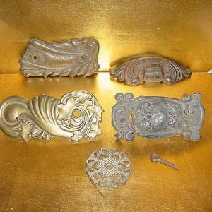 May include: An assortment of antique metal hardware pieces, including drawer pulls and decorative accents. The pieces are in various shapes and sizes, with ornate designs and a tarnished finish. The hardware is arranged on a gold-colored surface.
