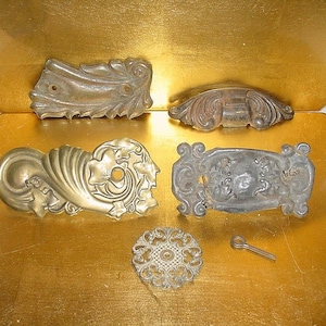 May include: An assortment of antique metal hardware pieces, including ornate drawer pulls and a decorative knob. The hardware features intricate designs and a tarnished finish, suggesting age and use. The pieces are arranged on a gold surface.