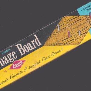 May include: A vintage Cribbage Board game box. The box is black and yellow with the words "Wood Cribbage Board" and "America's Favorite 2-handed Card Game!" printed on it. The game board is wood-toned with holes and pegs.