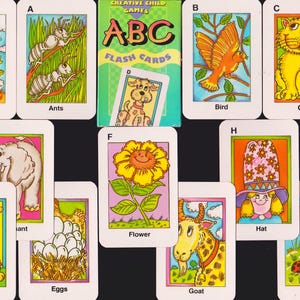May include: A collection of ABC flash cards featuring colorful illustrations of various objects and animals. The cards include images of airplanes, ants, a bird, a cat, a dog, eggs, a flower, a goat, a hat, and insects. The set comes in a green box.