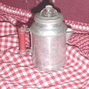 May include: Vintage aluminum coffee pot with a red wooden handle and a glass lid. The pot is sitting on a red and white checkered tablecloth. The coffee pot has a spout for pouring.