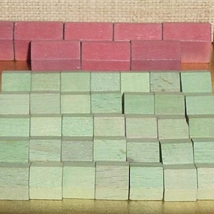 May include: A collection of small wooden game pieces shaped like houses, featuring a set of red house-shaped tokens arranged in the back and a larger group of light green square-shaped tokens arranged in a grid formation in the front.