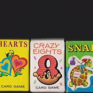 May include: Three vintage card game boxes: "Hearts" with heart graphics, "Crazy Eights" with a large number 8, and "Snap" featuring a turtle. Each box has a different color scheme and the words "Card Game".