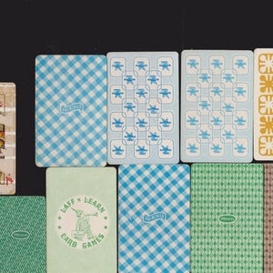 May include: A selection of vintage playing cards with diverse designs. The cards display patterns such as blue gingham, geometric shapes, and a green card with a cartoon character and the text "LAFF 'N LEARN CARD GAMES". Some cards show signs of wear.