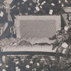 May include: A black and white photograph of a casket with a person inside. The casket is open and surrounded by floral arrangements. The interior of the casket is lined with a patterned fabric.