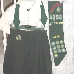 May include: A Girl Scout uniform set, including a white collared shirt with green accents, a dark green skirt, and a green sash. The sash features the number "1292" and various embroidered patches.