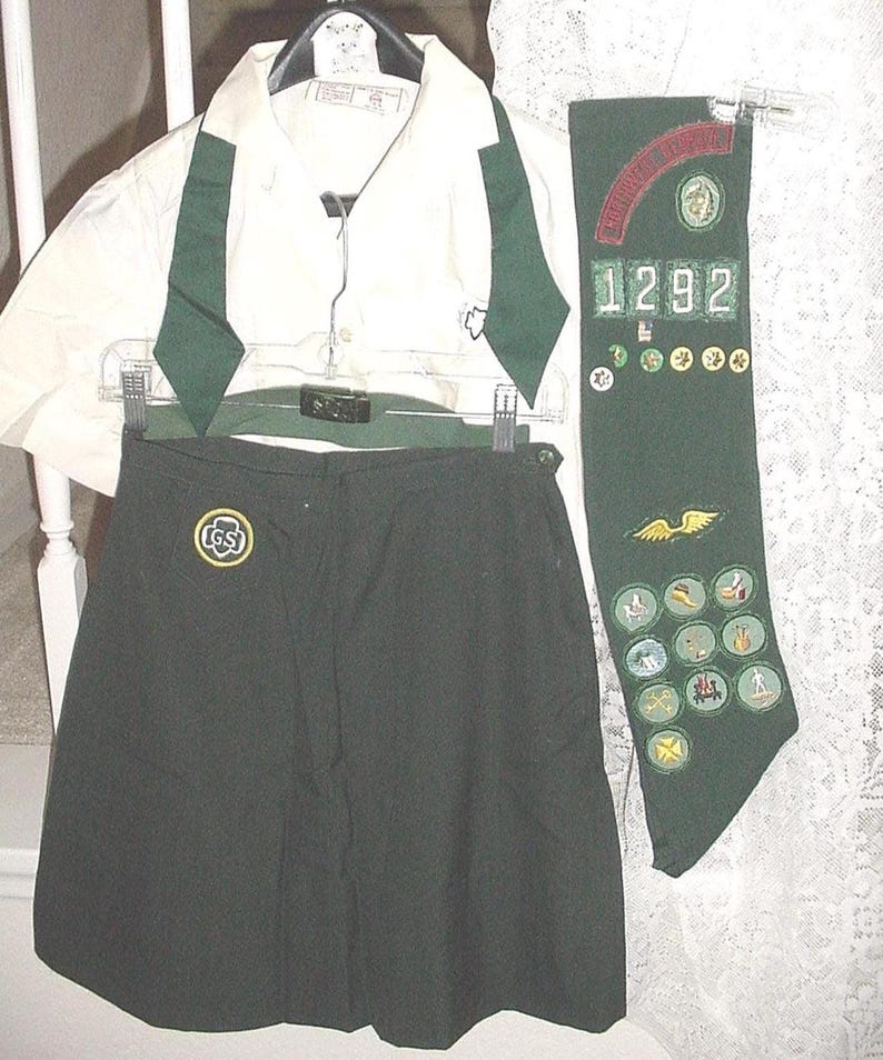 May include: A Girl Scout uniform set, including a white collared shirt with green accents, a dark green skirt, and a green sash. The sash features the number "1292" and various embroidered patches.