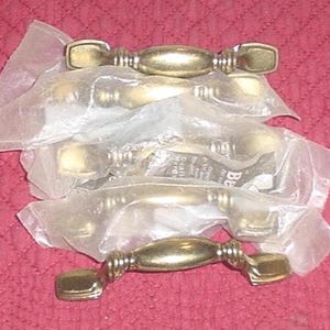 May include: A collection of brass-toned cabinet pulls, each in a clear plastic bag. The pulls have an oval center and rectangular ends, with decorative detailing. The hardware is designed for furniture and home improvement projects.