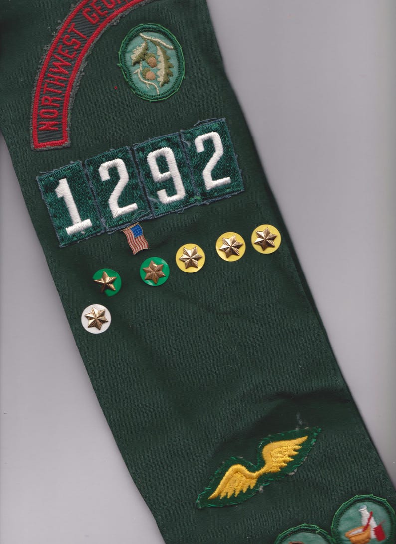 May include: A dark green sash with embroidered patches and pins. Patches include the text "NORTHWEST GEORGIA" in red, the number "1292" in white, and various emblems. Gold star pins and a small American flag pin are also visible.