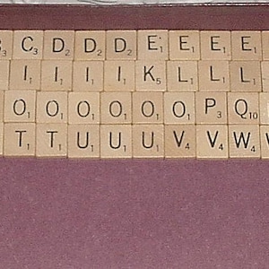 May include: A collection of light-colored wooden letter tiles arranged in rows within a dark purple container. Each square tile features a black capital letter and a small numerical subscript value, representing a standard set of game pieces.