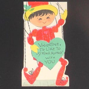 May include: Vintage Valentine's Day card featuring a puppet-like figure in red clothing and a yellow hat. The card has a green heart with the text "Valentine, I'd like to string along with you."