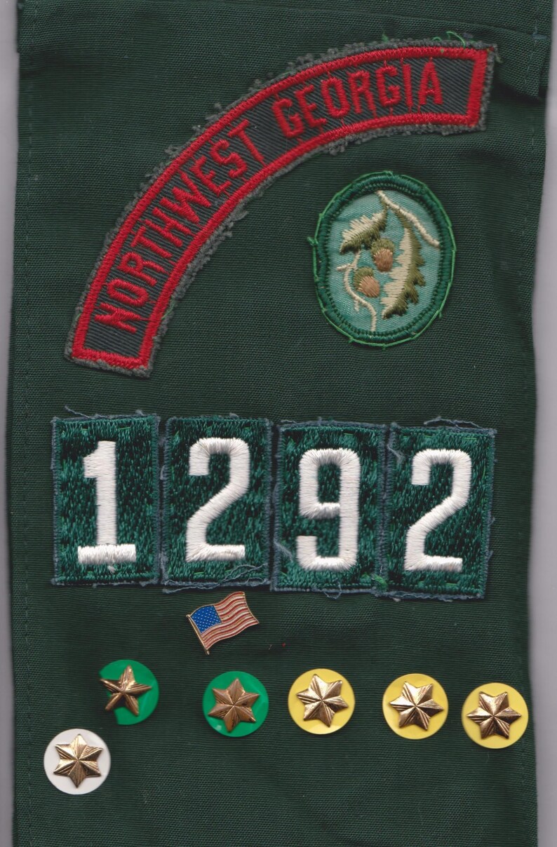 May include: Dark green fabric with embroidered patches and pins. Patches include "NORTHWEST GEORGIA" in red, a circular design, and the number "1292". Several star-shaped pins in gold and white are also present.