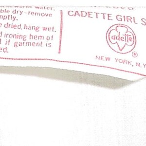 May include: A white fabric tag with red text and a red logo. The tag reads "CADETTE GIRL SCOUT" and includes washing instructions. The logo features the word "adette" inside a red emblem. The tag is from New York, N.Y.