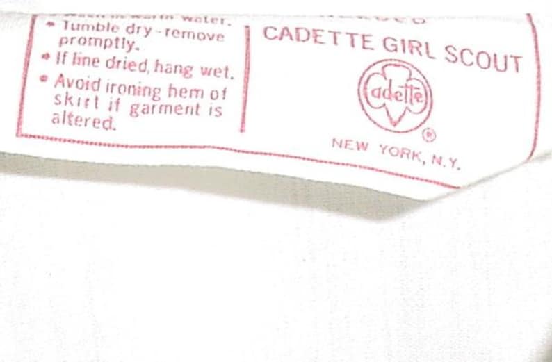 May include: A white fabric tag with red text and a red logo. The tag reads "CADETTE GIRL SCOUT" and includes washing instructions. The logo features the word "adette" inside a red emblem. The tag is from New York, N.Y.
