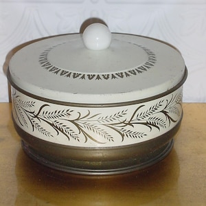 May include: A vintage white and gold metal container with a lid and a white knob. The container has a decorative leaf pattern around the sides and a geometric pattern on the lid. The container is sitting on a gold surface.