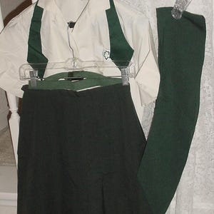May include: A cream-colored short-sleeved shirt with dark green accents and a small emblem, paired with a dark green skirt and leggings. The outfit is displayed on a hanger, suggesting a uniform or coordinated set.