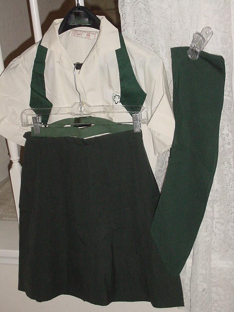 May include: A cream-colored short-sleeved shirt with dark green accents and a small emblem, paired with a dark green skirt and leggings. The outfit is displayed on a hanger, suggesting a uniform or coordinated set.