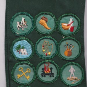 May include: A green fabric strip with embroidered patches featuring various designs. The patches include images of tools, musical instruments, and other symbols, with a yellow wing-like emblem at the top. The patches are circular and have a green border.