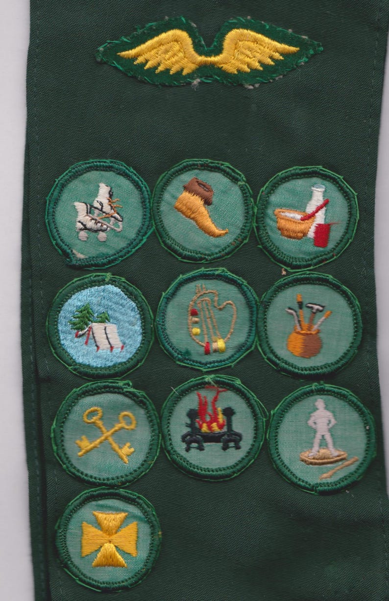 May include: A green fabric strip with embroidered patches featuring various designs. The patches include images of tools, musical instruments, and other symbols, with a yellow wing-like emblem at the top. The patches are circular and have a green border.
