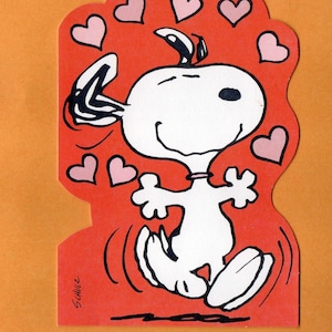 May include: A red die-cut card featuring a cartoon character with a white body, black ears, and a single black eye. The character is surrounded by pink hearts and appears to be dancing. The card has the signature "Schulz".