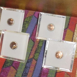 May include: Four square glass coasters with a copper-colored center. The coasters have a clear, beveled edge and a white interior. The coasters are displayed on a colorful, patterned surface.