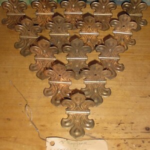 May include: A collection of fourteen metallic, copper-toned corner trim pieces, each featuring a symmetrical fleur-de-lis design with a central mounting hole. The pieces are arranged in a triangular formation, and a small paper tag reads Copper Corner Trim.