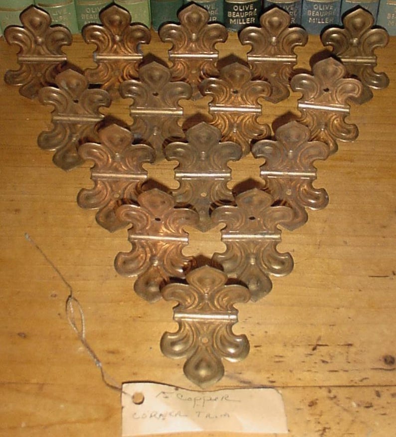 May include: A collection of fourteen metallic, copper-toned corner trim pieces, each featuring a symmetrical fleur-de-lis design with a central mounting hole. The pieces are arranged in a triangular formation, and a small paper tag reads Copper Corner Trim.