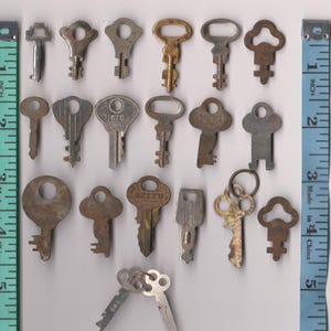 May include: A collection of twenty-two assorted metal keys of varying shapes, sizes, and finishes, arranged in rows between two vertical measuring tapes showing a length of approximately six inches for the entire display of keys.
