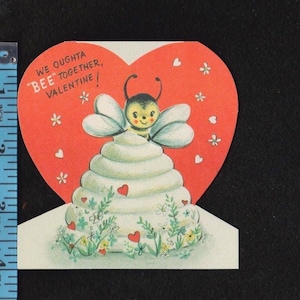 May include: A vintage Valentine's Day card with a bee theme. The card features a red heart shape with the text "We oughta BEE together, Valentine!" A white tiered structure is topped with a cartoon bee.