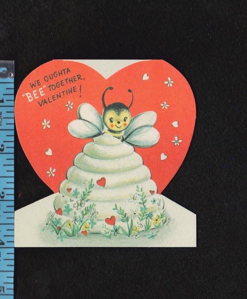 May include: A vintage Valentine's Day card with a bee theme. The card features a red heart shape with the text "We oughta BEE together, Valentine!" A white tiered structure is topped with a cartoon bee.