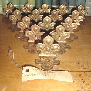 May include: A collection of fifteen metallic copper corner trim pieces, each featuring a fleur-de-lis design with a central hole for mounting. The pieces are arranged in a triangular formation, accompanied by a paper tag labeled 15 copper corner trim.