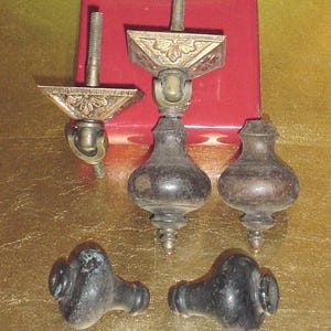 May include: A collection of five furniture hardware pieces including two brass-toned metal drawer pulls with ornate triangular backplates and threaded bolts, and three dark, rounded, bulbous-shaped knobs with a smooth, weathered finish.