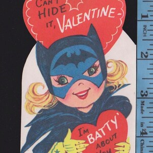 May include: A vintage-style paper Valentine card featuring a character in a blue bat mask and cape, holding a red heart that says 'I'M BATTY ABOUT YOU'. A red heart above says 'CAN'T HIDE IT, VALENTINE'. A ruler shows the card is 5 inches tall.