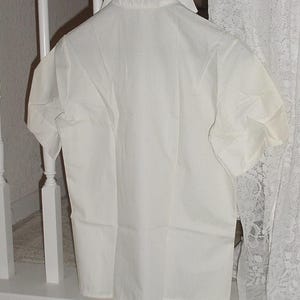 May include: A white short-sleeved shirt hanging on a metal hanger. The shirt has a collar and buttons down the front. The fabric appears to be cotton or a similar material. The shirt is hanging in front of a white railing and a lace curtain.