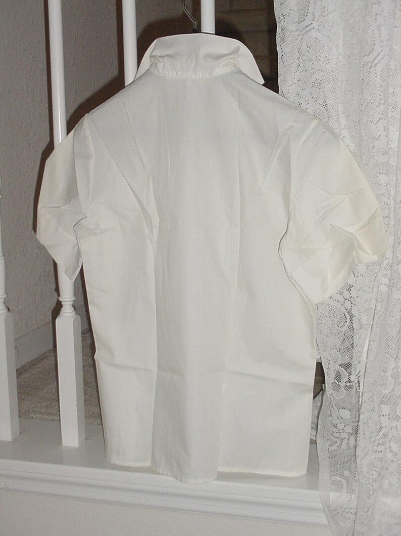May include: A white short-sleeved shirt hanging on a metal hanger. The shirt has a collar and buttons down the front. The fabric appears to be cotton or a similar material. The shirt is hanging in front of a white railing and a lace curtain.