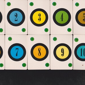 May include: Ten playing cards with white backgrounds, each featuring a number from 1 to 10. The numbers are inside a colored circle, with a black border. The colors are orange, blue, yellow, green, and brown. Each card has a green dot in each corner.