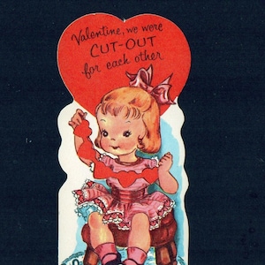 May include: Vintage Valentine's Day card featuring a cartoon girl holding a heart-shaped cutout. The card has a red heart-shaped top with the text "Valentine, we were CUT-OUT for each other." The girl is wearing a pink dress and sitting on a small stool.