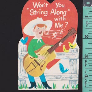 May include: A heart-shaped paper valentine card featuring a person in a white cowboy hat playing a brown acoustic guitar, with the text "Won't You String Along with Me?" and a measuring tape showing a height of approximately 5 inches.