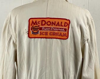 Può includere: Cappotto vintage della divisa McDonald's color crema. Il cappotto presenta una cintura in vita e una toppa sul retro con il logo McDonald's e la scritta "Super Creamed Ice Cream". Il cappotto è a maniche lunghe e abbottonato.