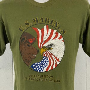 May include: Olive green t-shirt featuring a circular graphic with a bear and an eagle facing each other, separated by a US flag. The text "U.S. MARINES" arches above the graphic, with the phrase "DEFEND FREEDOM OR LEARN TO SPEAK RUSSIAN" below.