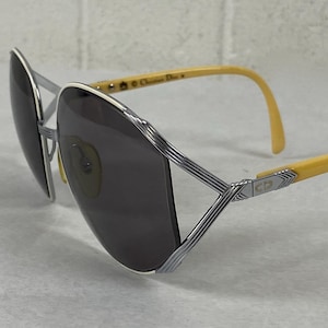 May include: Vintage sunglasses with a silver frame and gray lenses. The temples are yellow, and the design includes the initials "CD". The sunglasses are set against a neutral background.