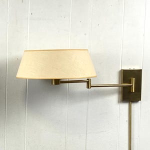 May include: A wall-mounted lamp featuring a beige lampshade and a brass-coloured adjustable arm. The lamp is mounted on a rectangular brass-coloured base. The lamp is plugged into an electrical outlet.