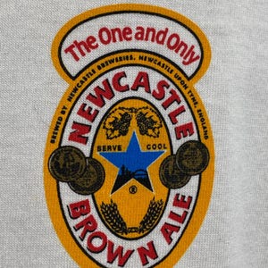 May include: A close-up of a white t-shirt featuring a Newcastle Brown Ale logo. The logo is a circular design with the words "Newcastle Brown Ale" in red and black, with a blue star and wheat stalks in the center.