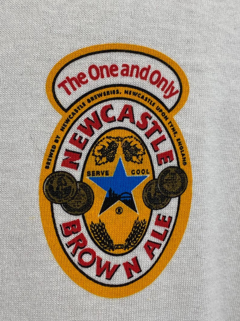 May include: A close-up of a white t-shirt featuring a Newcastle Brown Ale logo. The logo is a circular design with the words "Newcastle Brown Ale" in red and black, with a blue star and wheat stalks in the center.