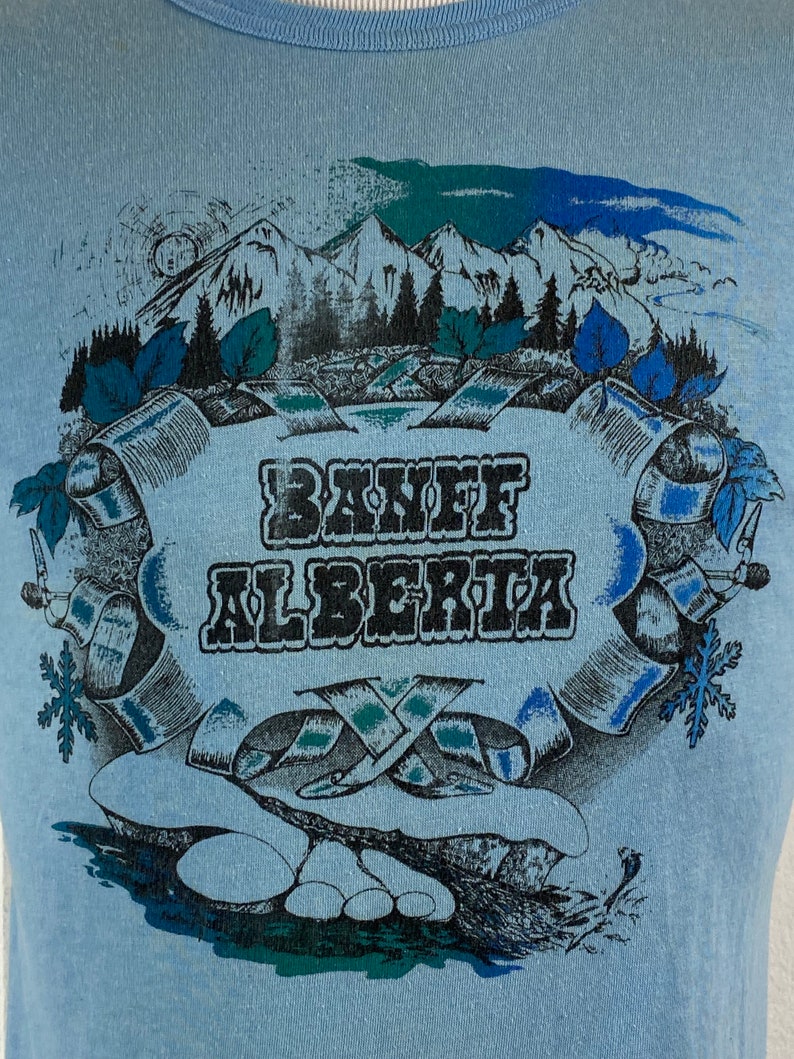 K&ouml;nnte beinhalten: Ein Vintage-T-Shirt in Blau mit einem grafischen Aufdruck einer Bergszene und dem Text "Banff Alberta".