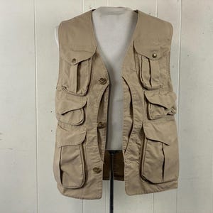 May include: A tan utility vest with multiple pockets of varying sizes. The vest has a button-up front and a V-neck design. The vest is made of a durable fabric and is designed for outdoor activities or work.