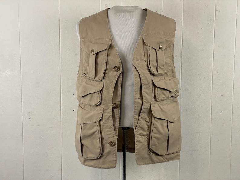 May include: A tan utility vest with multiple pockets of varying sizes. The vest has a button-up front and a V-neck design. The vest is made of a durable fabric and is designed for outdoor activities or work.