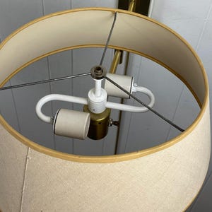 May include: A close-up of a beige lampshade with a gold trim. The lamp has a brass-coloured base and a white light fixture. The lamp is designed to be mounted on a wall. The lamp is a vintage style.