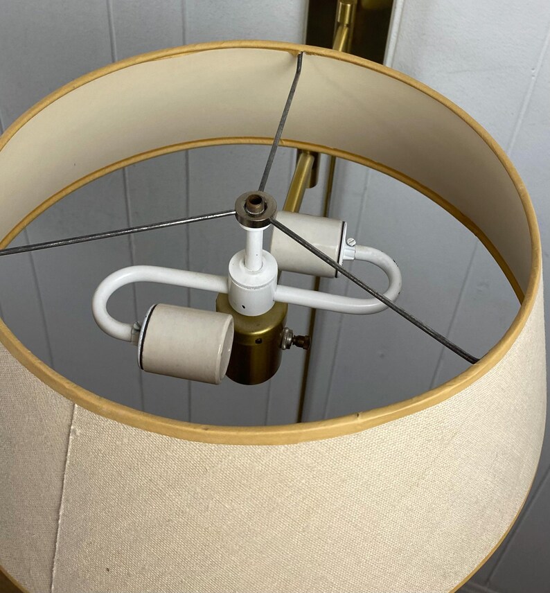 May include: A close-up of a beige lampshade with a gold trim. The lamp has a brass-coloured base and a white light fixture. The lamp is designed to be mounted on a wall. The lamp is a vintage style.